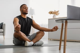 Mann meditiert im Wohnzimmer auf Matte: Meditieren fördert die Konzentration und hilft bei der Stressbewältigung. Dadurch wird die Achtsamkeit im Alltag verbessert.