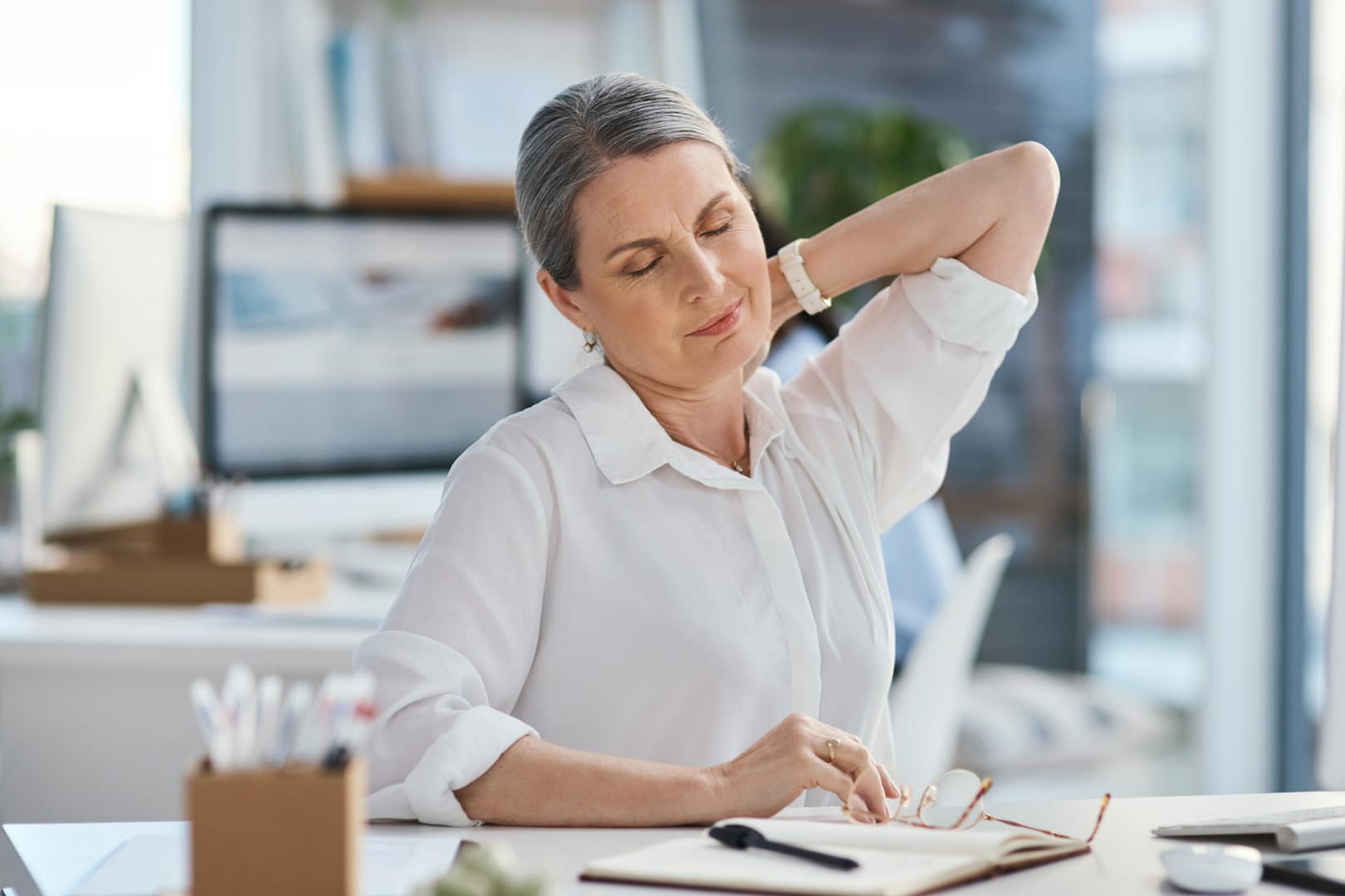 Älter Frau mit Nackenschmerzen im Büro: Eine ungünstige Haltung, wie es häufig bei Schreibtischarbeit der Fall ist, kann sich auf Dauer stark auf Rücken und Nacken auswirken.