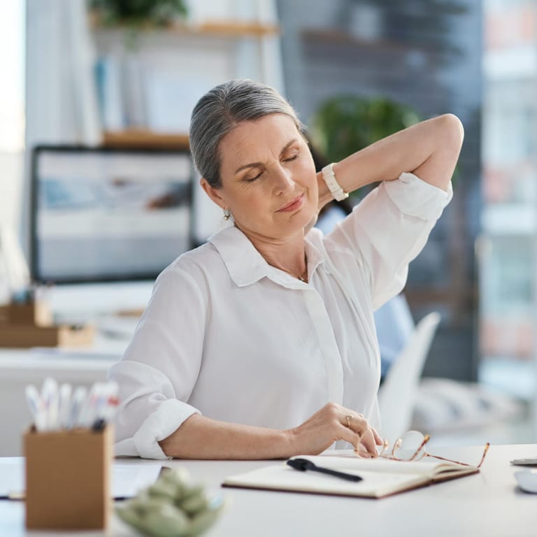 Älter Frau mit Nackenschmerzen im Büro: Eine ungünstige Haltung, wie es häufig bei Schreibtischarbeit der Fall ist, kann sich auf Dauer stark auf Rücken und Nacken auswirken.