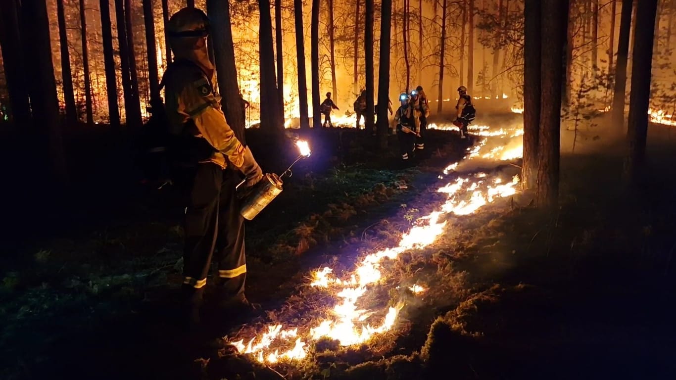 Auf dem flachen Boden wird ein Feuer entzündet, im Hintergrund lodert schon der große Waldbrand.