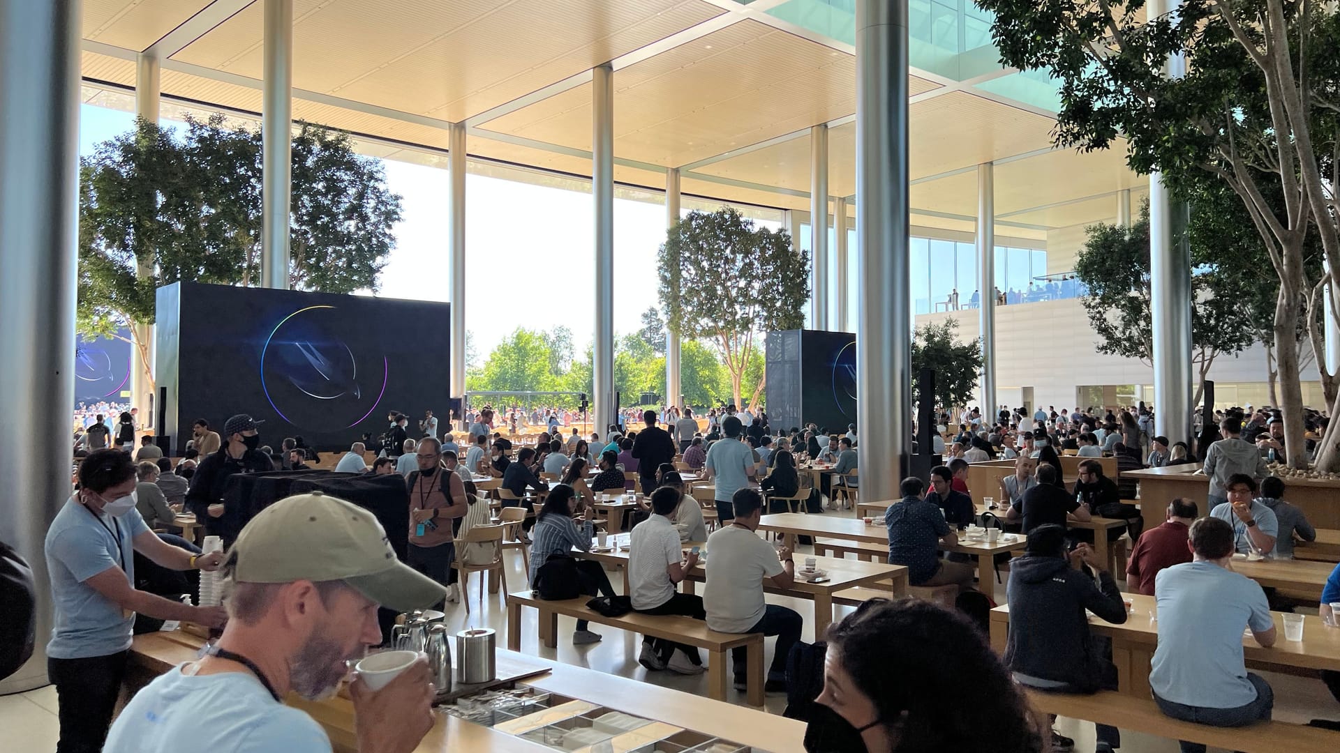 Ein Blick in einen Cafeteria-Bereich. Die Glasfassade ist hier über Dutzende Meter zur Seite geschoben worden. Etliche Stuhlreihen setzen sich nach draußen außerhalb des Rings in den Park fort, wo für die Keynote eine riesige Leinwand aufgebaut wurde.