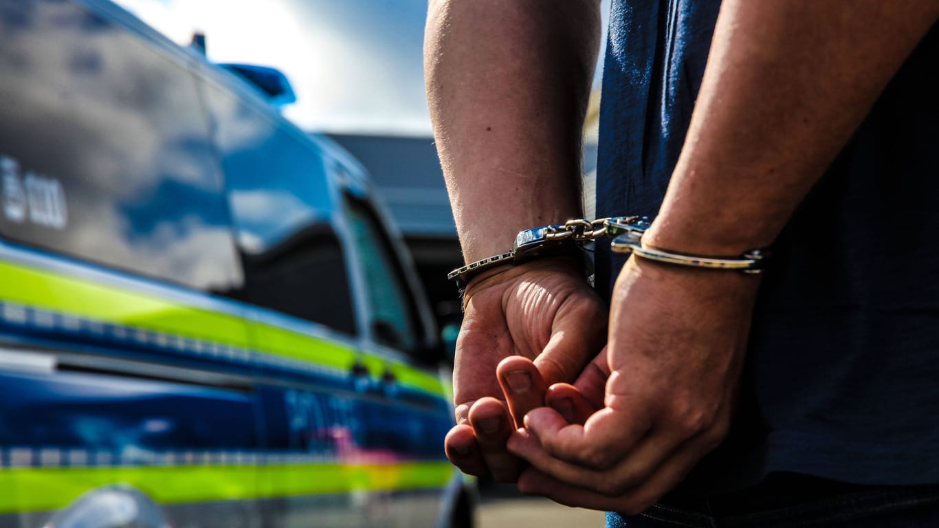 Eine Person ist mit Handschellen gefesselt und steht vor einem Streifenwagen (Symbolbild). Die beiden Verdächtigen wurden vorläufig festgenommen.