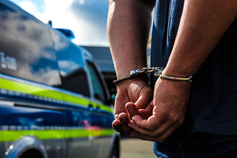 Eine Person ist mit Handschellen gefesselt und steht vor einem Streifenwagen (Symbolbild). Die beiden Verdächtigen wurden vorläufig festgenommen.