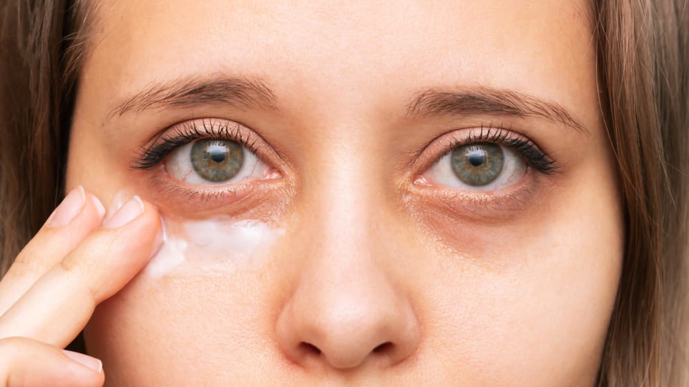 Frau trägt Augencreme auf Augenringe auf: Augencremes versorgen die Haut unter den Augen mit Feuchtigkeit und haben einen glättenden Effekt. Die untere Augenpartie wirkt dadurch voller und die dunklen Schatten lassen sich besser abdecken.