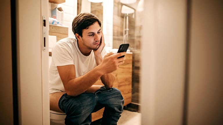 Ein Mann sitzt auf Toilette und schaut auf sein Smartphone.