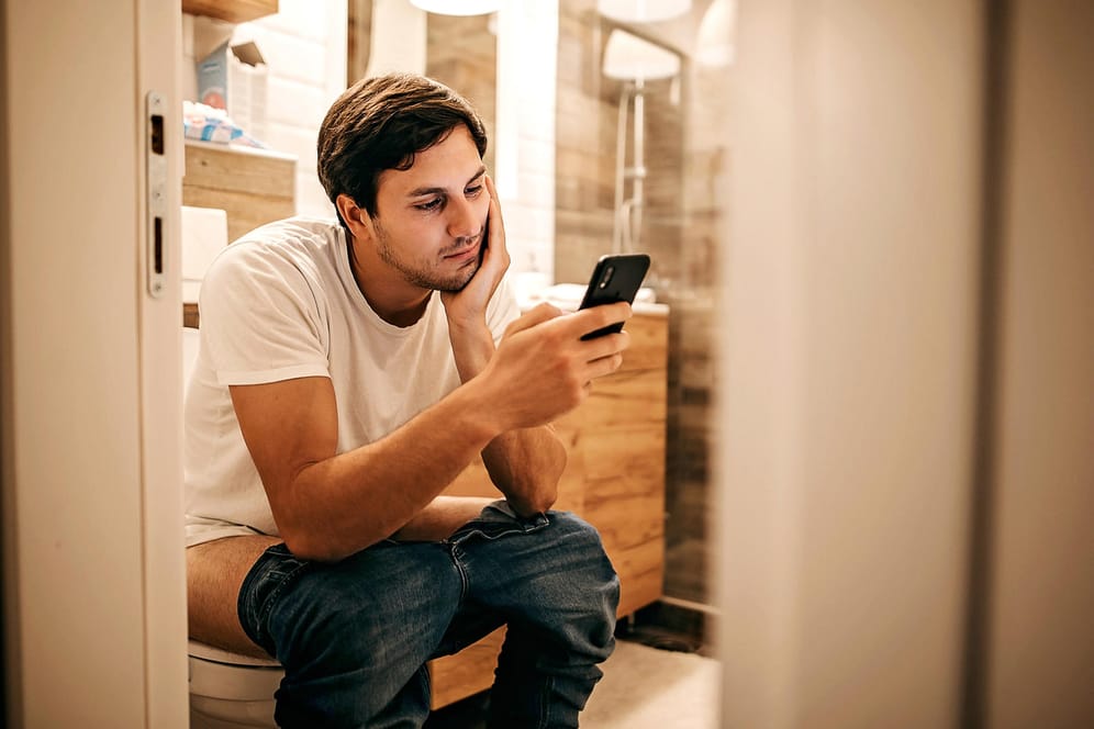 Ein Mann sitzt auf Toilette und schaut auf sein Smartphone.