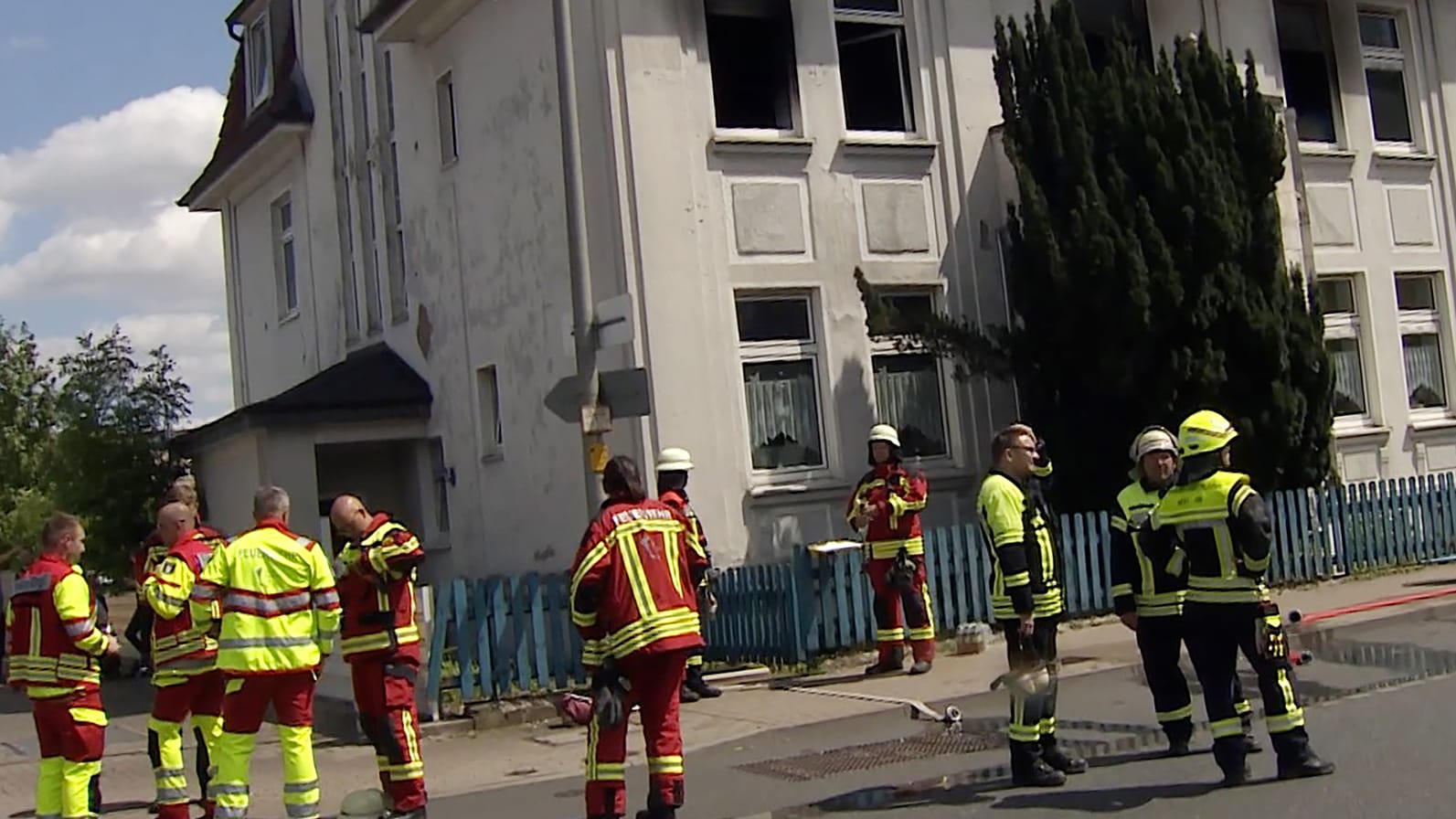 Verrußte und beschädigte Fenster und sind an dem Mehrfamilienhaus zu sehen. Davor stehen Feuerwehrleute.