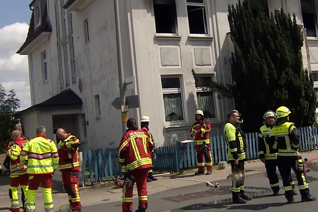 Verrußte und beschädigte Fenster und sind an dem Mehrfamilienhaus zu sehen. Davor stehen Feuerwehrleute.