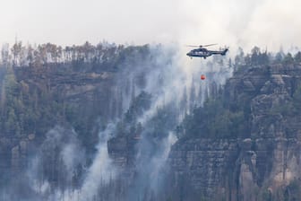Sachsen, Schmilka: Ein Hubschrauber fliegt mit einem Löschwasser-Außenlastbehälter über einen Brand im Nationalpark Sächsische Schweiz.