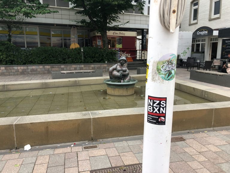 Ein Fahnenmast mit Aufklebern vor der Wilhelmine: Der eine Sticker macht Werbung für einen Cannabis-Community, der andere mit dem Schriftzug "NZS BXN" (Nazis boxen) ruft zum gewaltsamen Antifaschismus auf.