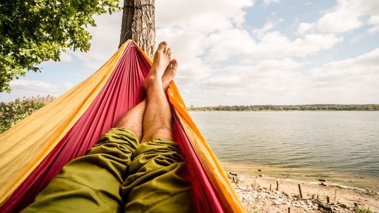 Relaxing in the hammock at the beach under a tree, summer day. Barefoot man laying in hammock, looking on a lake, inspiring landscape