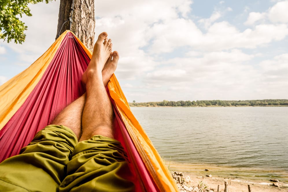 Relaxing in the hammock at the beach under a tree, summer day. Barefoot man laying in hammock, looking on a lake, inspiring landscape