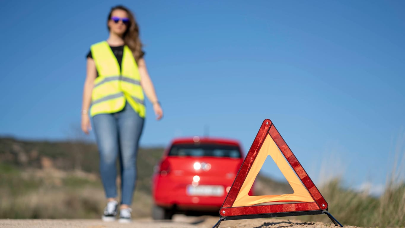 Ein Auto steht am Straßenrand. Eine Frau mit Warnweste steht daneben. Im Vordergrund ist ein Sicherheitsdreieck.