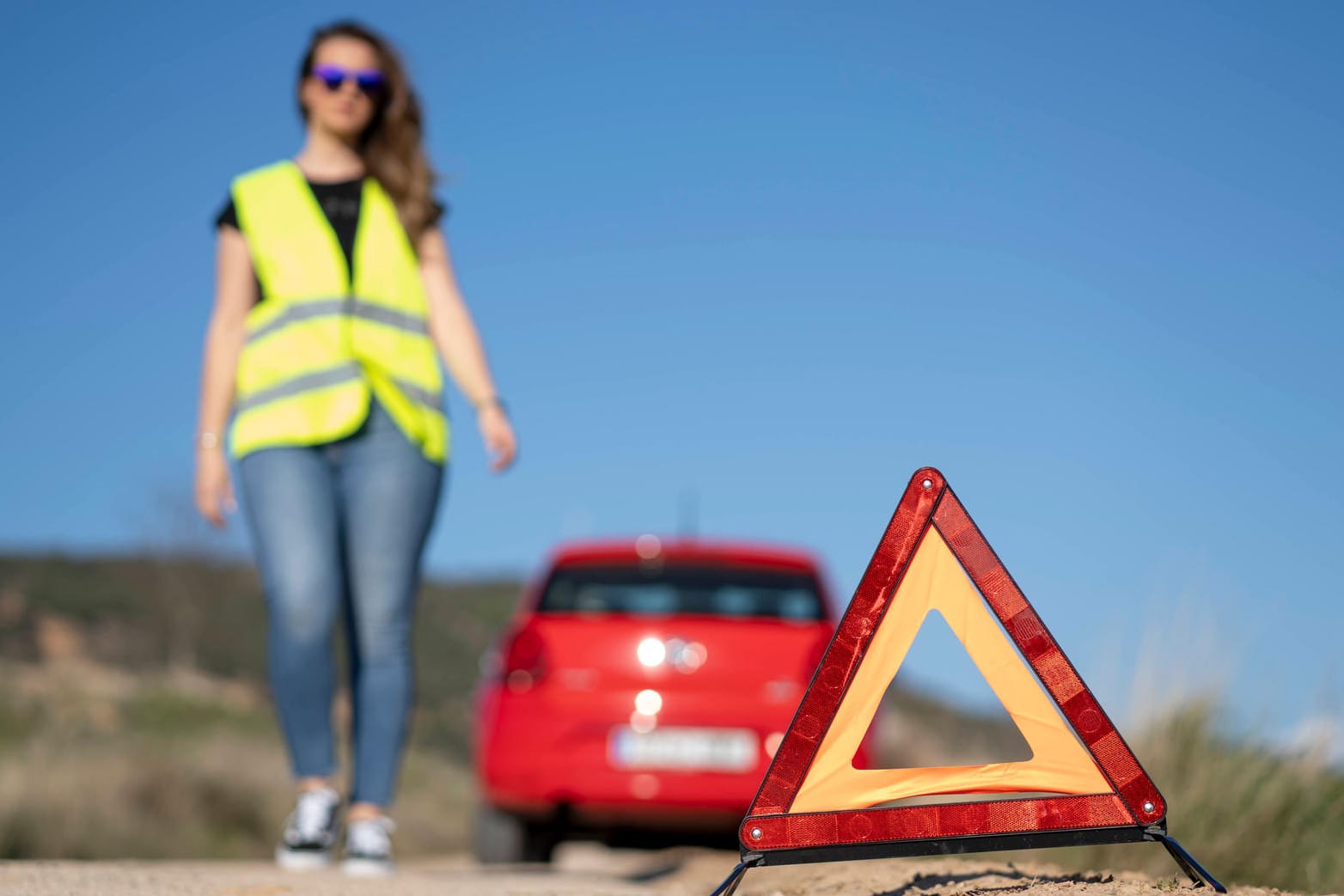 Ein Auto steht am Straßenrand. Eine Frau mit Warnweste steht daneben. Im Vordergrund ist ein Sicherheitsdreieck.