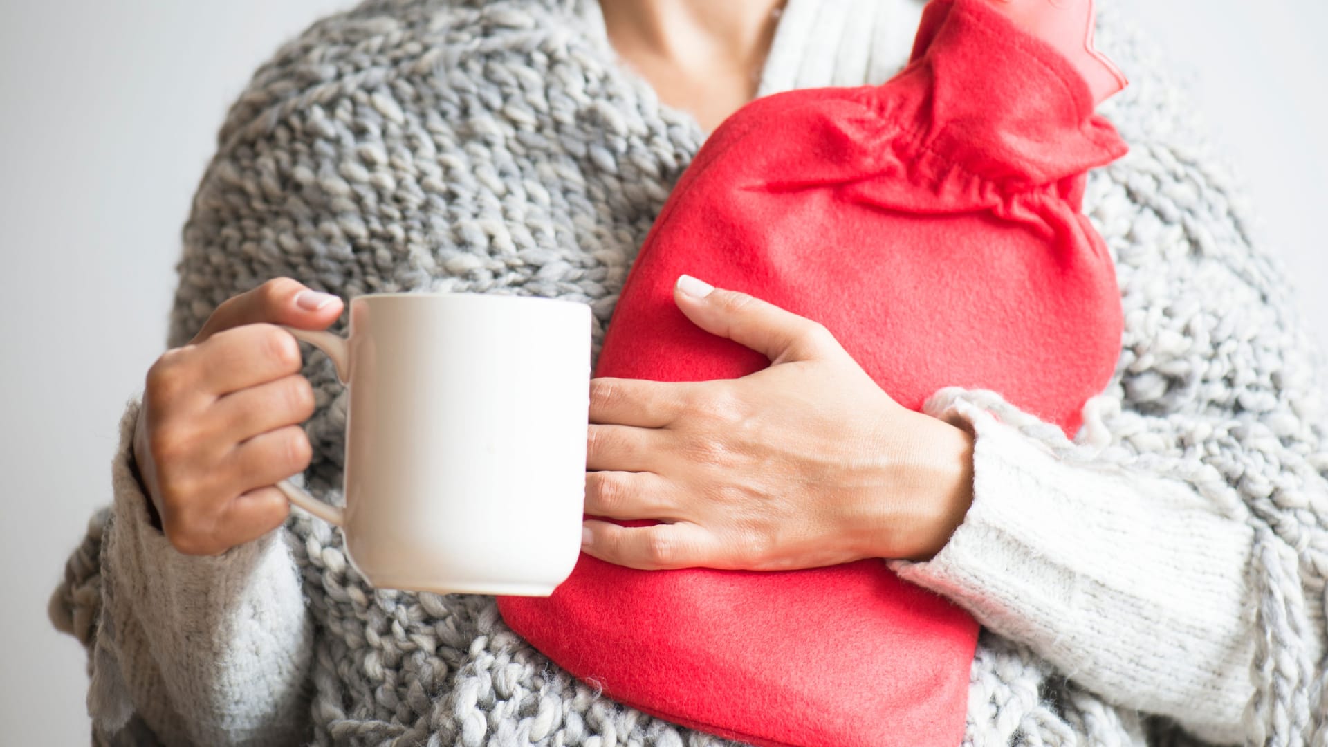 Eine Frau hält eine Wärmflasche im Arm und eine Tasse in der Hand.