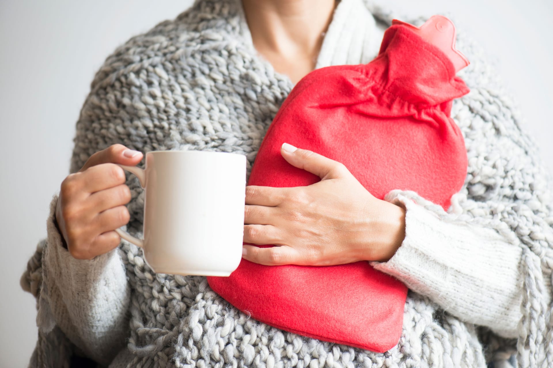 Eine Frau hält eine Wärmflasche im Arm und eine Tasse in der Hand.