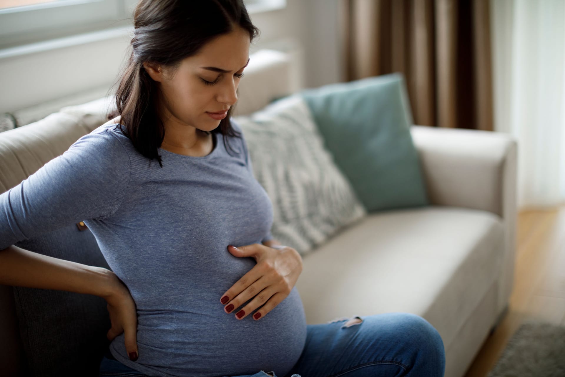 Eine schwangere Frau sitzt auf dem Sofa und fasst sich mit einer Hand an den unteren Rücken und mit der anderen an den Bauch.)