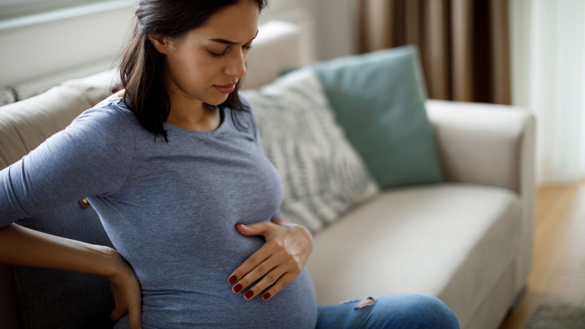 Eine schwangere Frau sitzt auf dem Sofa und fasst sich mit einer Hand an den unteren Rücken und mit der anderen an den Bauch.)