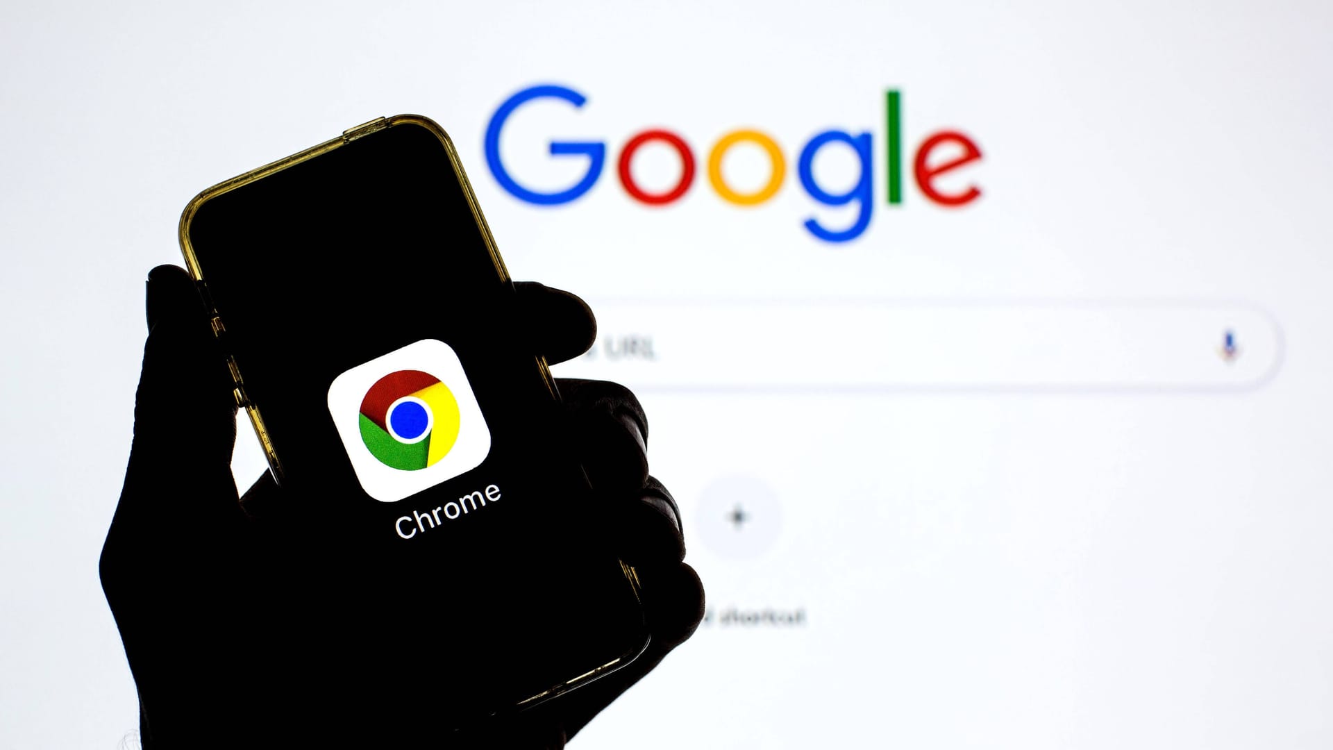 Google-Suchleiste wird auf einem Bildschirm gezeigt und davor wird ein Handy mit dem Google-Chrome-Logo gezeigt.