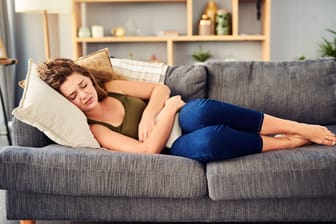 Eine Frau liegt auf dem Sofa und hält sich eine Wärmflasche an den Bauch.