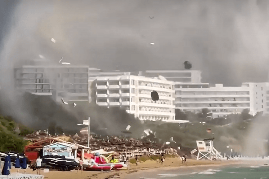 Eine Windhose fegt über einen Badestrand und wirbelt Schirme und Liegen in die Luft.