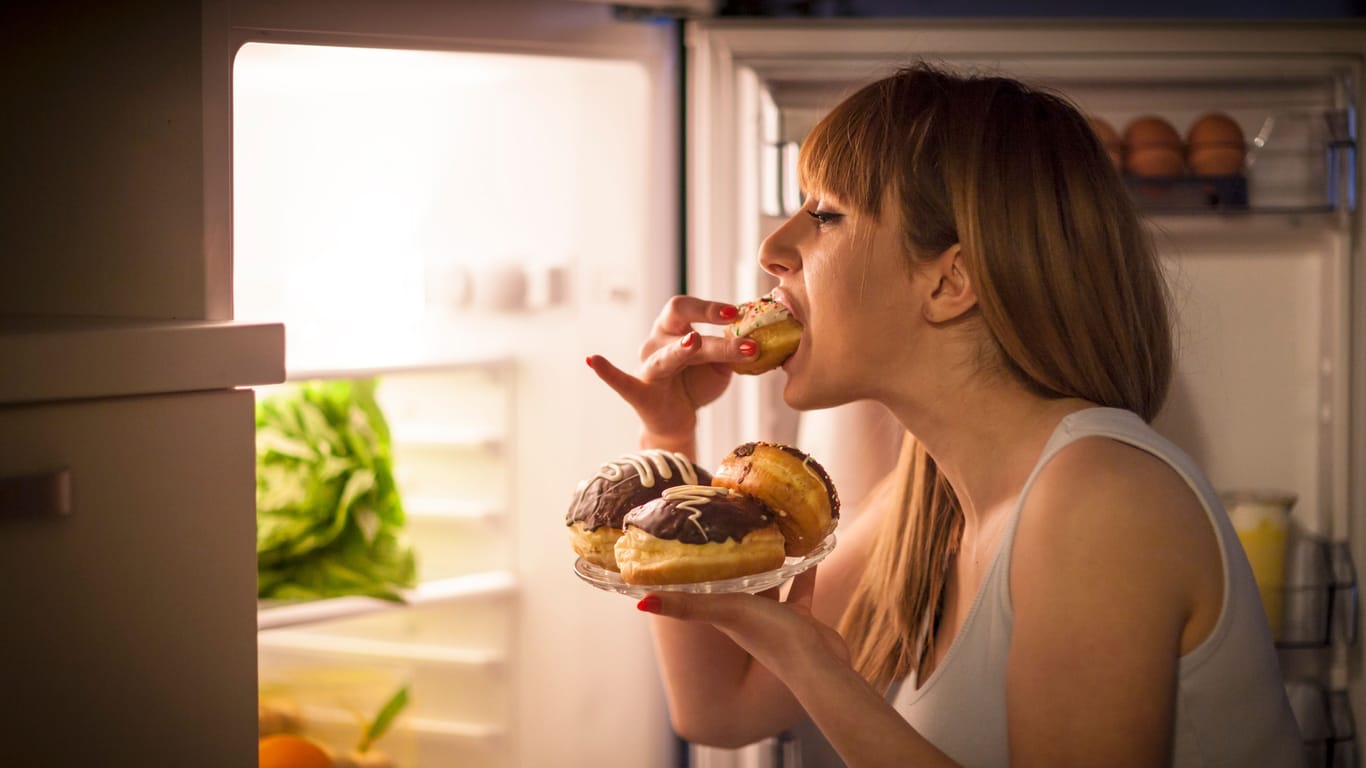 Eine Frau steht vor dem geöffneten Kühlschrank und isst Gebäck.