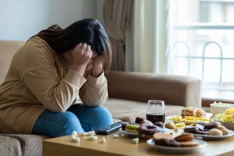 Eine Frau sitzt vor einem Tisch, auf dem viel ungesundes Essen steht, und sieht sehr traurig aus.