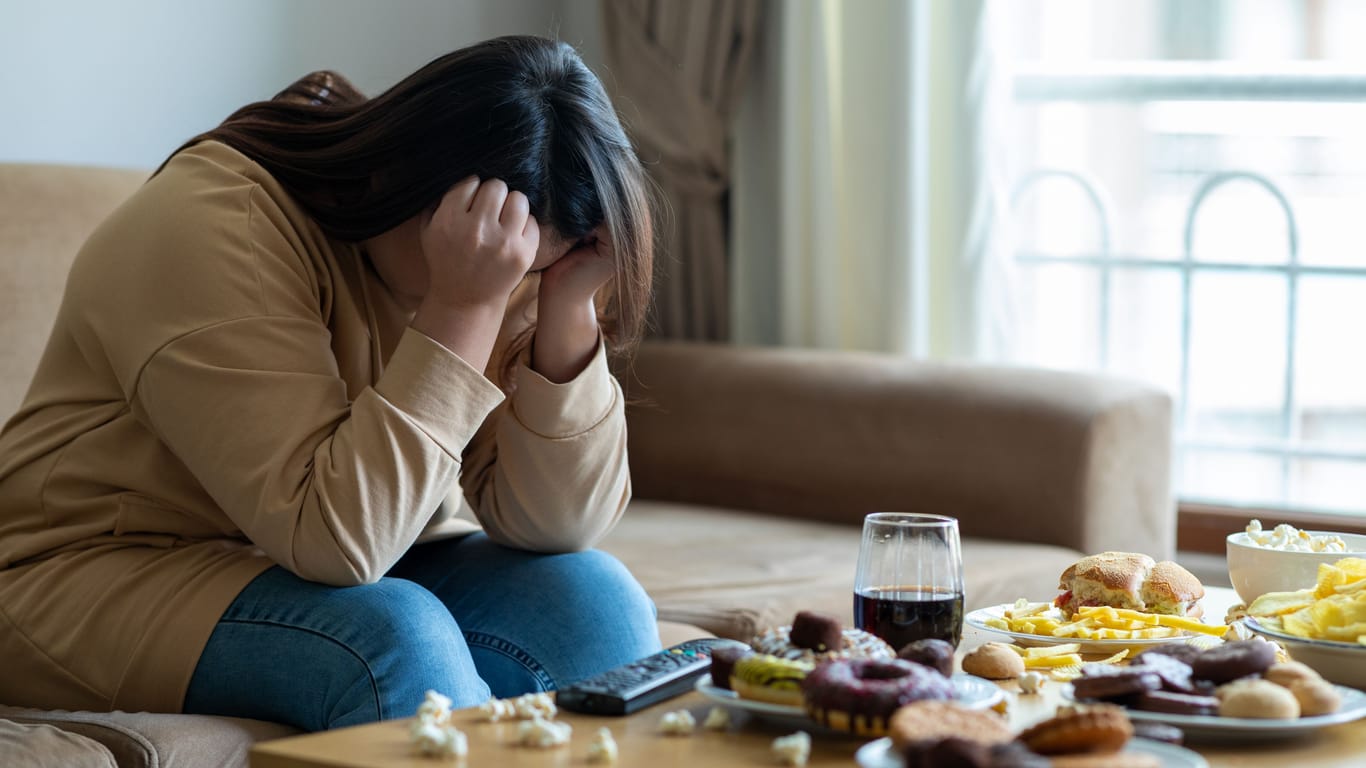 Eine Frau sitzt vor einem Tisch, auf dem viel ungesundes Essen steht, und sieht sehr traurig aus.