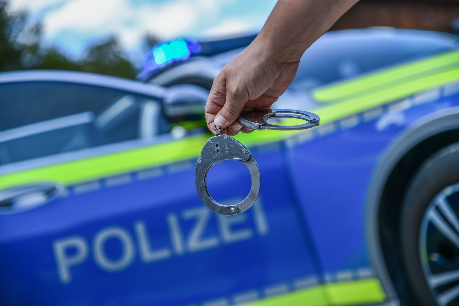 Eine Person hält Handschellen vor einem Dienstwagen der Polizei (Symbolbild): Ein Verdächtiger konnte ermittelt werden.