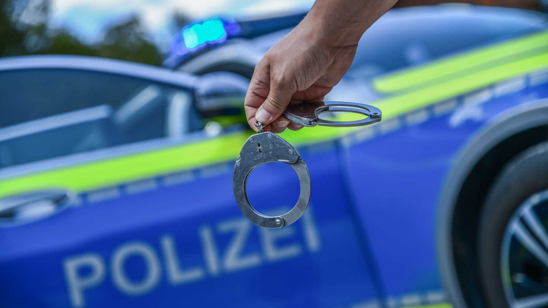 Eine Person hält Handschellen vor einem Dienstwagen der Polizei (Symbolbild): Ein Verdächtiger konnte ermittelt werden.