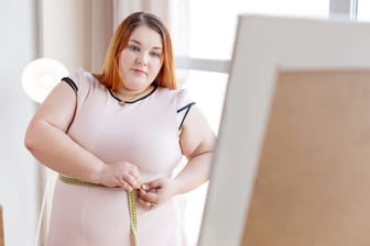 Eine übergewichtige Frau steht vor dem Spiegel und legt ein Maßband um ihren Bauch.