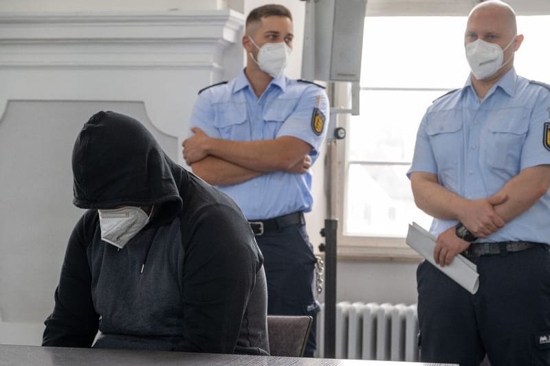 Der Angeklagte sitzt im Landgericht an einem Tisch und wird von Justizbeamten bewacht.