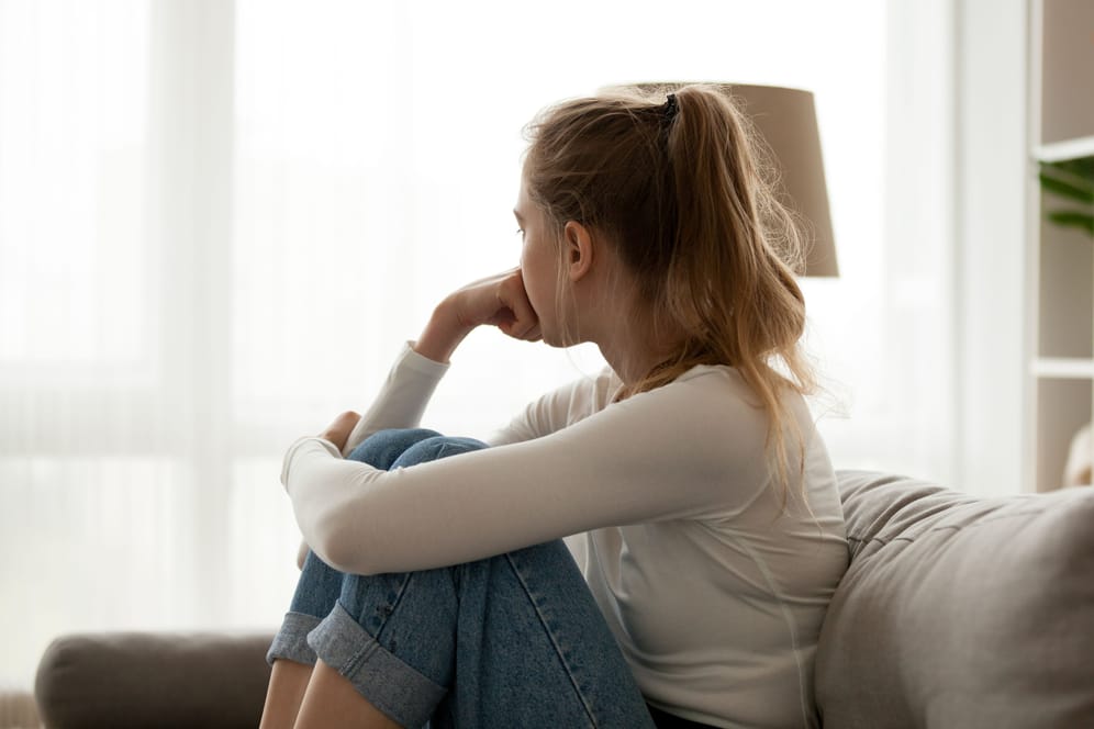 Ein Mädchen sitzt auf dem Sofa und schaut traurig aus dem Fenster.