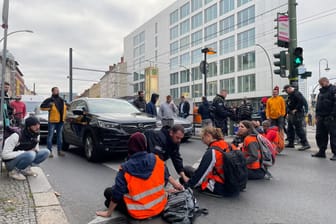 Ein Polizist löst festgeklebte Aktivisten in Berlin von der Straße ab (Archivbild): Rund eine halbe Stunde Zeitverzögerung gibt es aktuell.