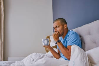 Mann sitzt krank im Bett und hustet