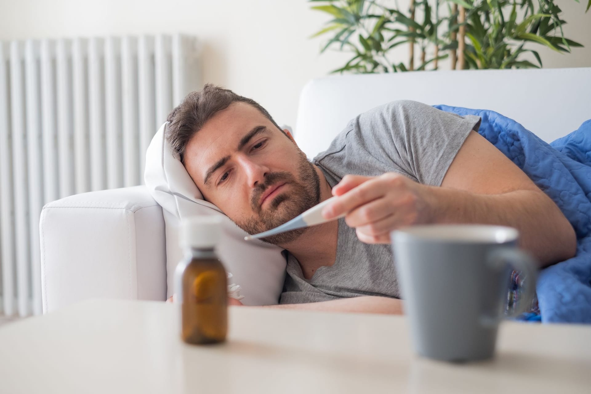 Ein Mann liegt auf dem Sofa und schaut auf ein Fieberthermometer.