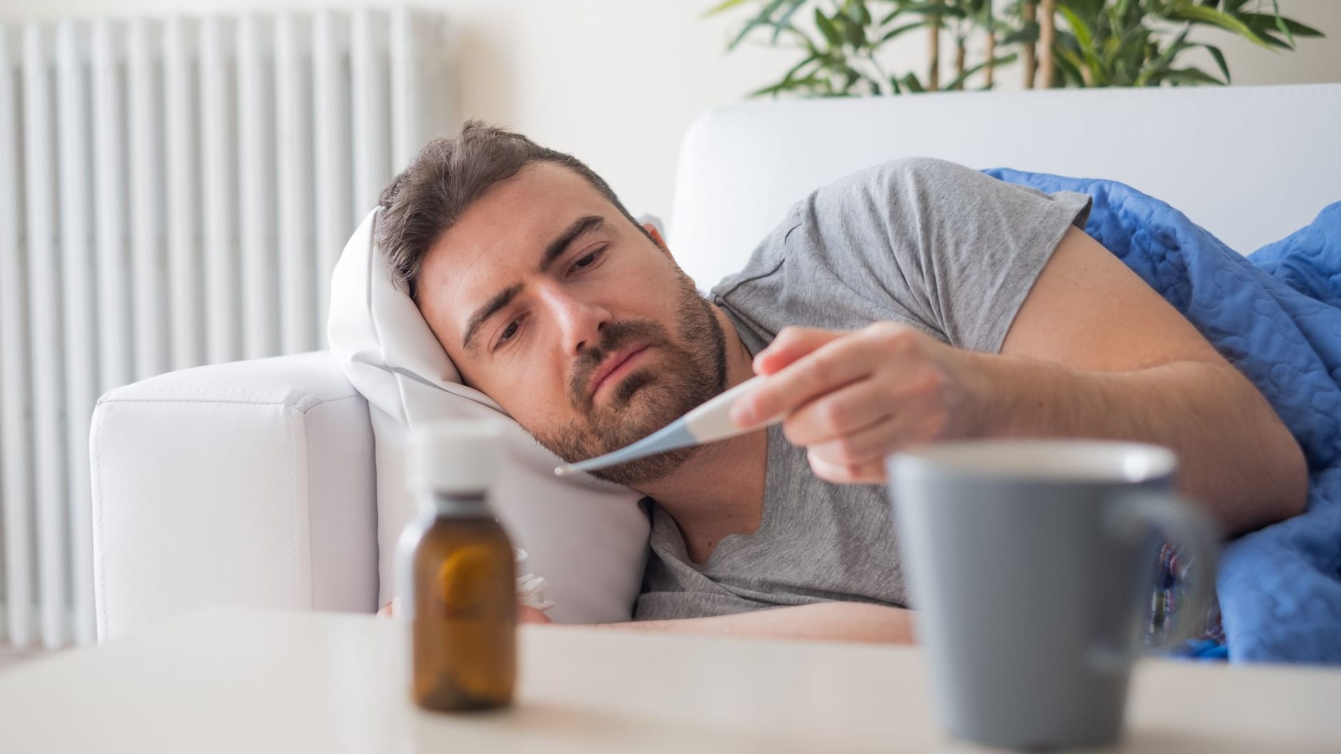 Ein Mann liegt auf dem Sofa und schaut auf ein Fieberthermometer.