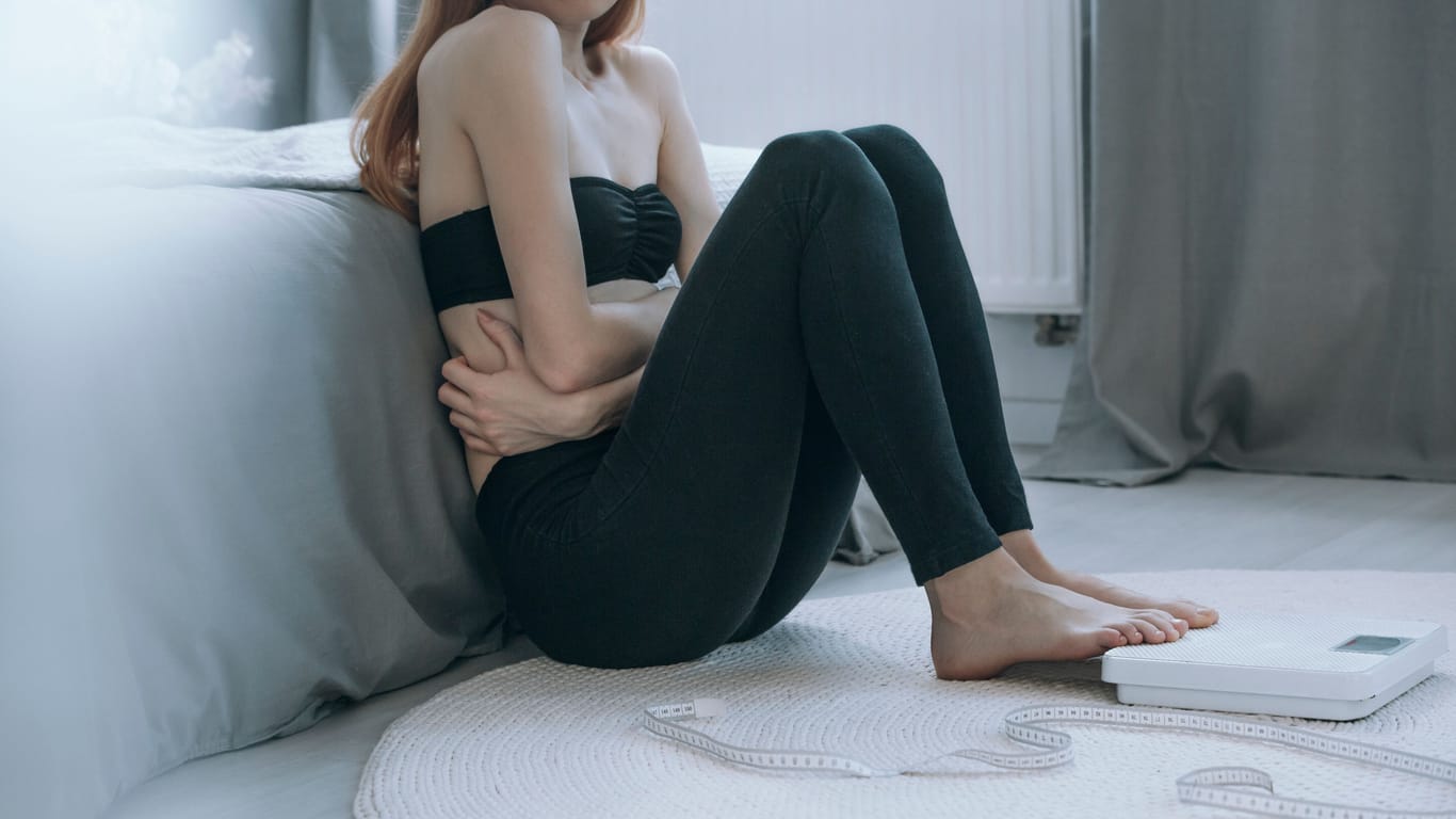 Ein stark untergewichtiges Mädchen sitzt auf dem Boden und hält sich den Bauch.