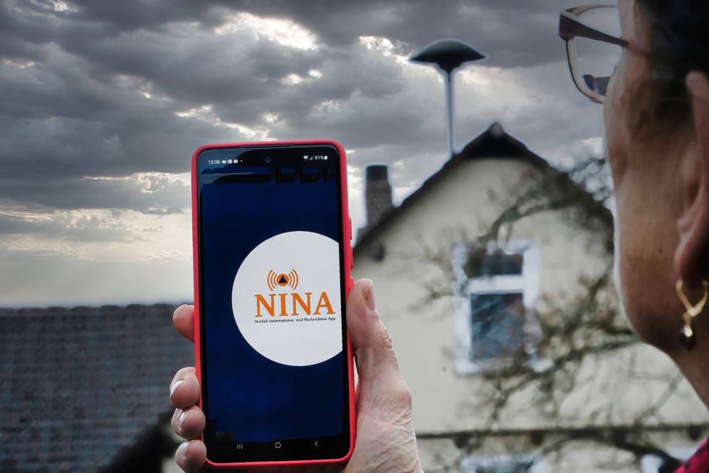 Eine Frau blickt auf eine Sirene und hat die Warn-App Nina auf ihrem Handy geöffnet (Symbolbild): Am Donnerstag geht der Warntag in die zweite Runde.
