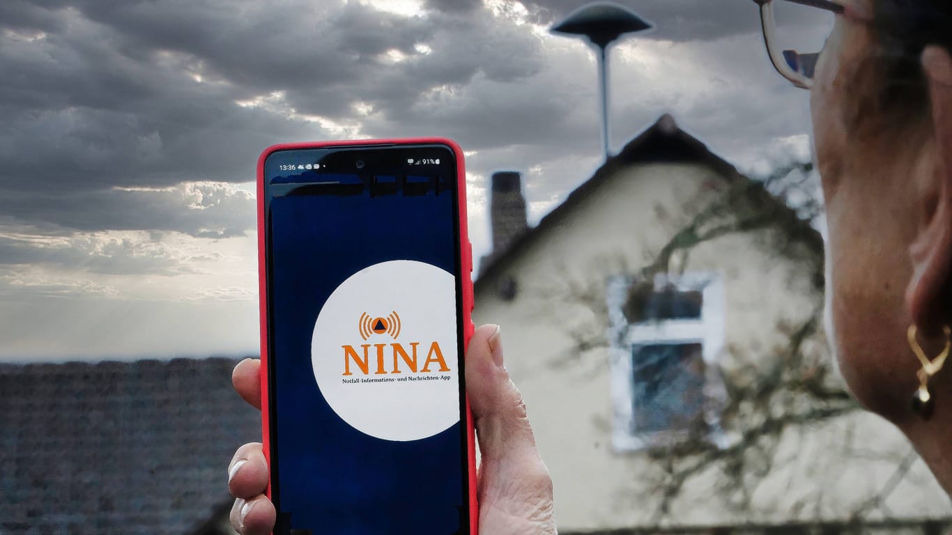 Eine Frau blickt auf eine Sirene und hat die Warn-App Nina auf ihrem Handy geöffnet (Symbolbild): Am Donnerstag geht der Warntag in die zweite Runde.