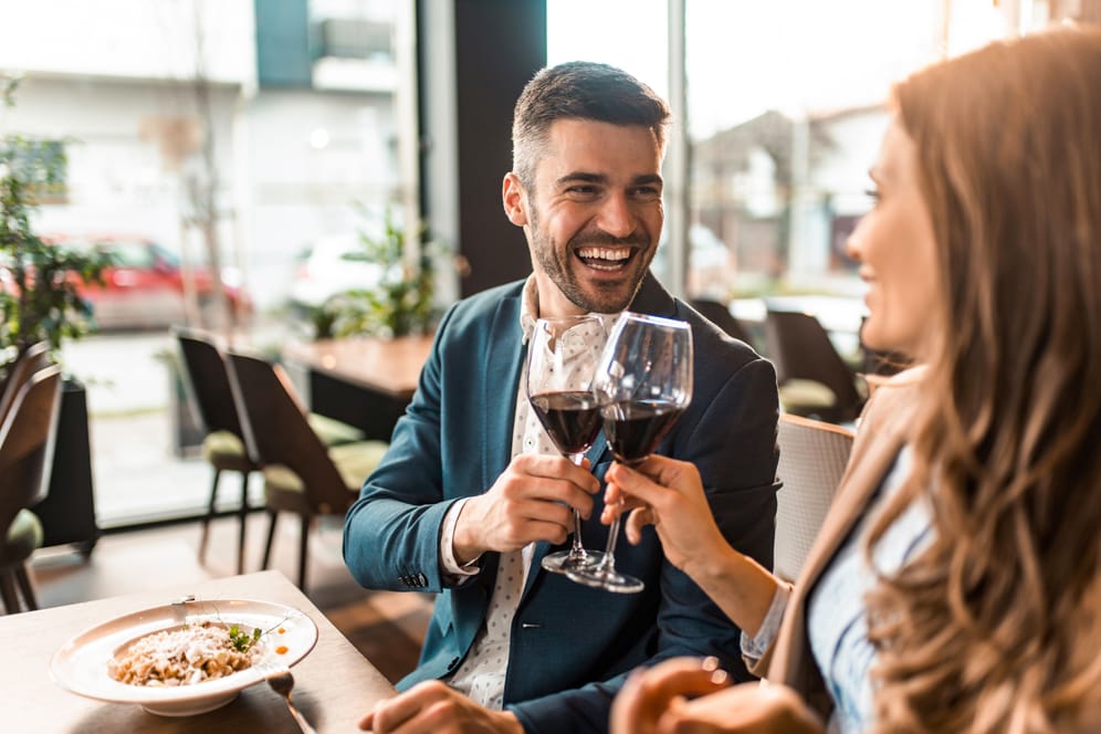 Eine Frau und ein Mann stoßen beim Essen mit einem Glas Rotwein an.