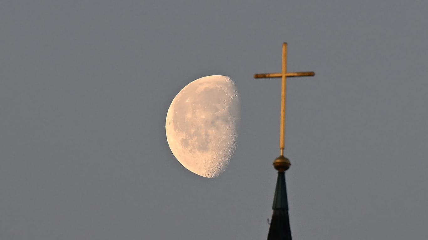 Der Mond steht im Morgenlicht hinter einem Kreuz auf einem Kirchturm (Symbolbild): In Berlin muss der Kirchenaustritt beim Amtsgericht erklärt werden.
