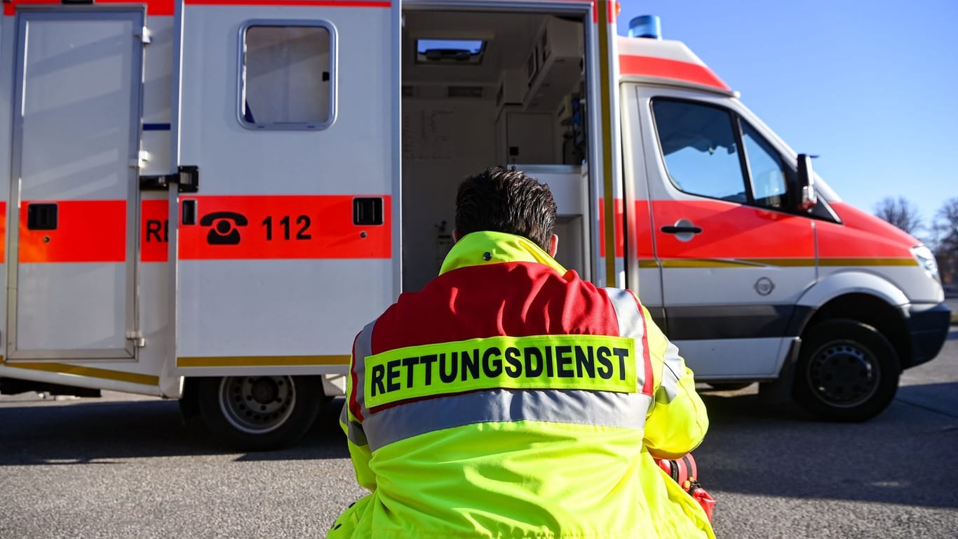 "Rettungsdienst" steht auf Jacke eines Mannes vor einem Rettungswagen (Symbolbild): Rettungskräfte beklagen Personalmangel und warnen vor dem Kollaps.