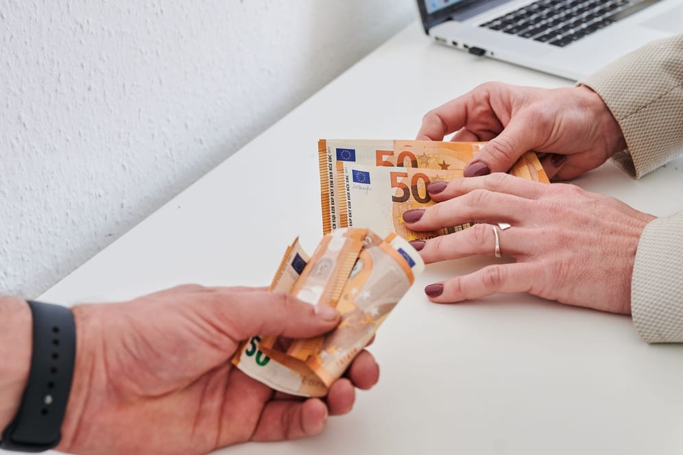 Eine Männerhand hält ein Bündel 50-Euro-Scheine, während eine Frauenhand nur wenige 50-Euro-Scheine hält (Symbolbild): Frauen verdienen in NRW pro Stunde weniger als Männer.