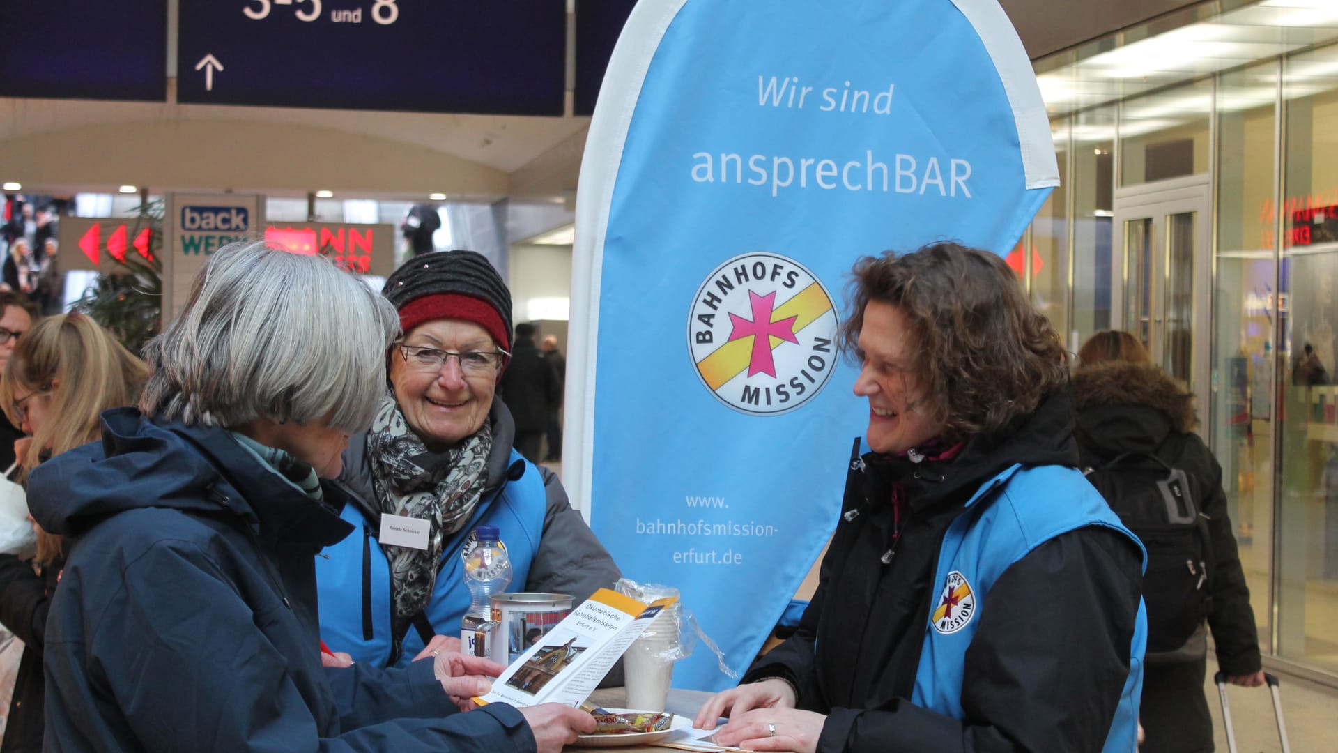 Zwei Ehrenamtliche der Bahnhofmission sprechen an ihrem Stand mit einer Frau