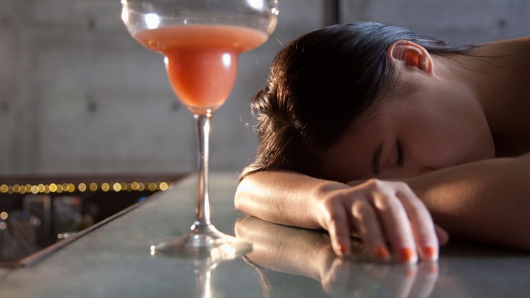 Eine Frau liegt mit dem Kopf auf der Bar, vor ihr steht ein Cocktail.