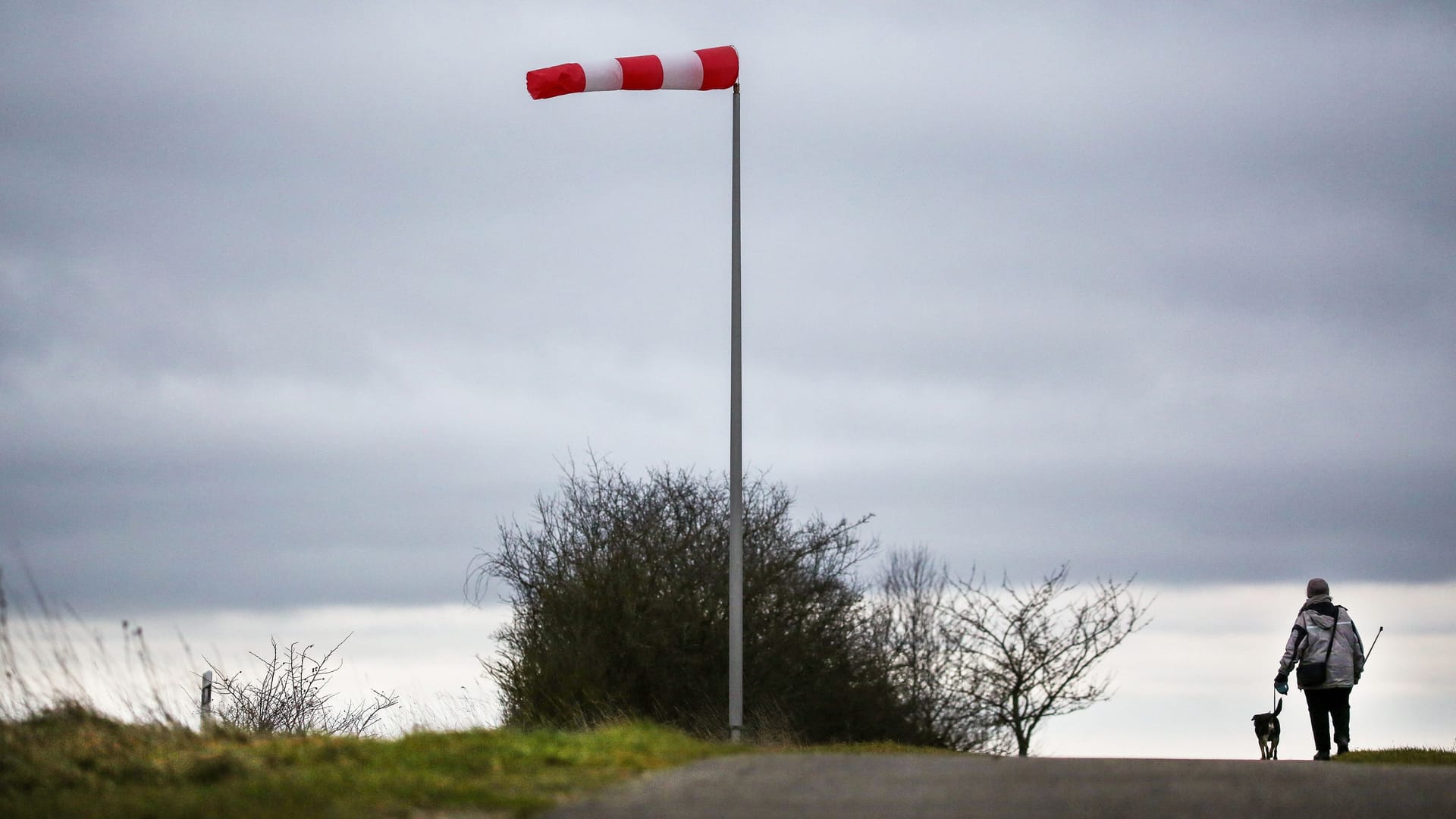 Eine Frau geht bei stürmischem Wind mit ihrem Hund an einem Windsack vorbei (Archivbild): In NRW ist stürmisches Wetter angekündigt.