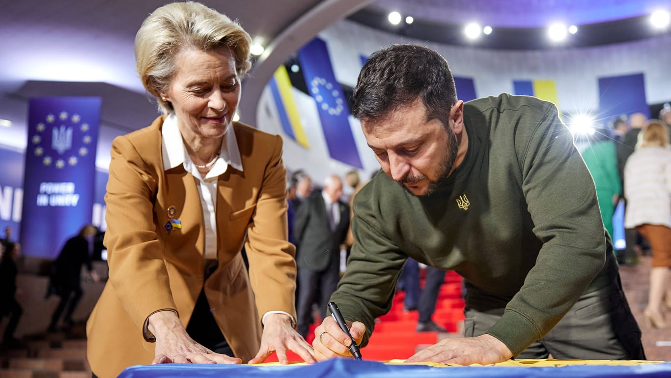 Ukraine-Gipfel: EU-Kommissionspräsidentin Ursula von der Leyen strafft die ukrainische Flagge, während der Präsident des Landes, Wolodymyr Selenskyj, darauf unterschreibt.