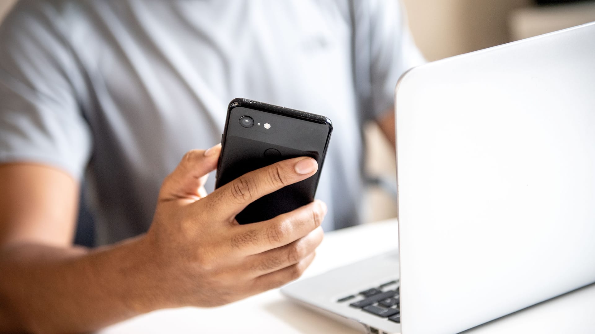 Ein junger Mann sitzt mit Smartphone vorm Laptop (Symbolbild): Steuern auf dem Smartphone erklären – mit "MeinELSTER+" soll es ganz einfach möglich sein.