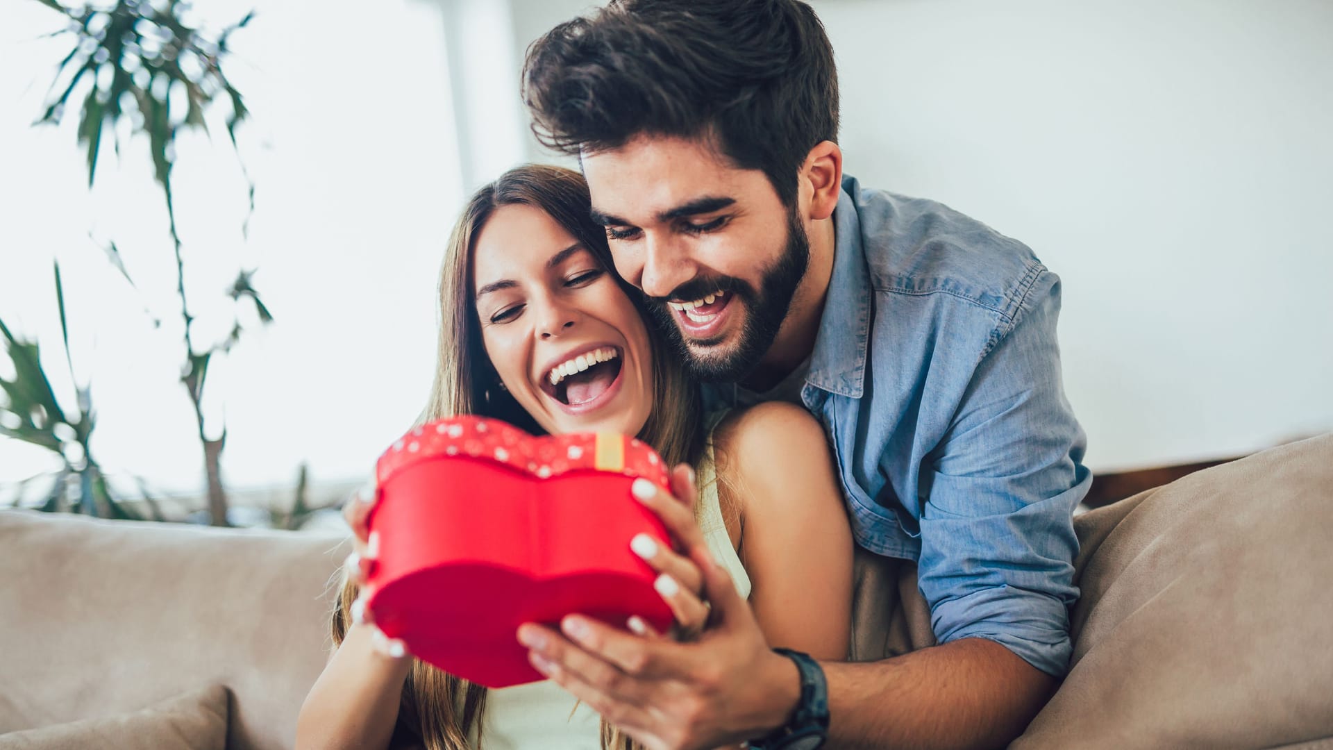 Beautiful young couple is celebrating at home. Handsome man is giving his girlfriend a gift box